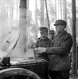 During additional refresher training, a Finnish soldier has his breakfast served into a mess kit by another soldier from a steaming field kitchen in the forests of the Karelian Isthmus. More soldiers, two of them visible, wait in line for their turn behind him. It is early October, and the snow has not yet set in.