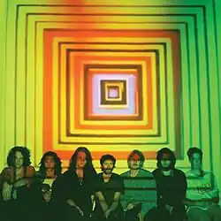 The members of King Gizzard sit in a line against a wall, while a pattern of squares is projected on the wall behind them.