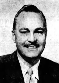 Black-and-white portrait photograph of a suited, smiling man with a pencil mustache. His dark hair is slicked back in a pompadour, with no sideburns.