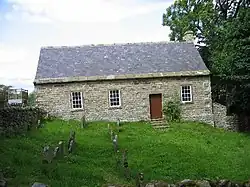 A small, stone, single-storey building with a slate roof in a burial ground with gravestones. Seen from the front, it has steps leading up to a door and three windows.