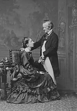 A couple is shown: On the left is a tall woman of about 30. She wears a voluminous dress and is sitting sideways in an upright chair, facing and looking up into the eyes of the man who is on the right. He is about 60, quite short, and balding at the temples. He is dressed in a suit with a tailcoat and wears a cravat. He faces and looks down at the woman. His hand rests on the back of the chair.
