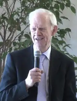An elderly man in a suit speaking into a microphone with a tree in the background