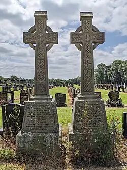 Two tall beige stone celtic cross gravestones of Geoffrey Burke and Frederick Johnson with respective family members, with smaller, squarer graves and green grass visible in the background