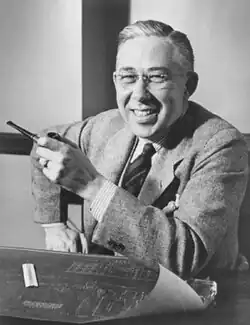 A photograph of Stoddard at his desk in front of a blueprint, gesturing with a pipe.