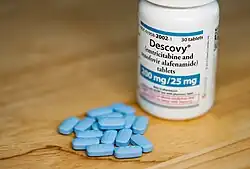 A photo of a small white medicine bottle standing next to a small collection of small sky-blue pills on a wooden table. The pills have a distinct beveled shape.