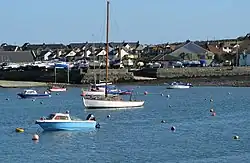 Foreground: A handful of tethered boats floating on calm water, surrounded by small buoys. Background: Raised shoreline of rock and concrete, topped by boats lined up beside each other; in the centre a slipway meets the water. In the far background is a collection of small residential buildings.