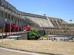 Ground-level view of a large concrete structure in the left, power lines at the top and a parking lot with many cars in it