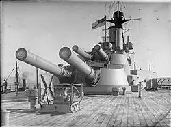 Two large gun turrets seen from the deck of a battleship; each turret has two long guns