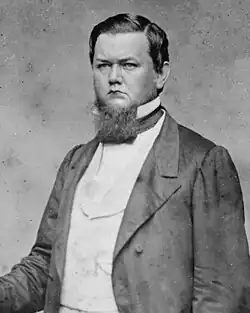 A man in his thirties with black hair and a black beard. He is wearing a white shirt and black jacket.