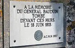 A color photograph of a plaque. The text in all caps and French reads "A la memoire General Bauduin tombe devant ces murs. Le 18 Juin 1815. (Logo carved) A.C.M.N 1957".