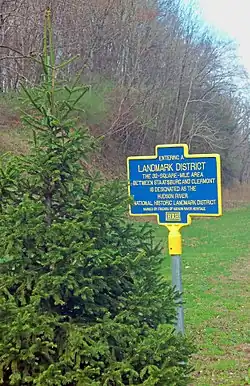 A sign with the headline "Landmark District" in yellow on a blue background, with yellow trim, behind an evergreen tree on its left. There are some trees in back.
