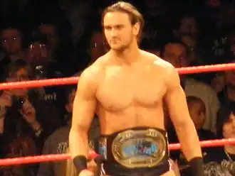 A Caucasian male stands in a wrestling ring with red ropes. He has long dark hair tied back, and is wearing short, black wrestling tights. A championship belt is around his waist, with a black strap and gold center plate.
