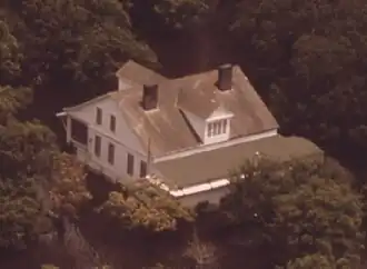 A white house with prominent dormers on the roof surrounded by trees which are flooded and surrounded by water.