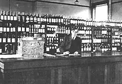 A photo of a man behind the counter at a liquor store with shelves stocked with liquor behind him.