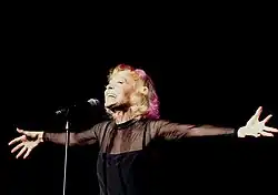 A smiling woman with short blond hair faces a microphone. Her arms are flung wide open to either side.