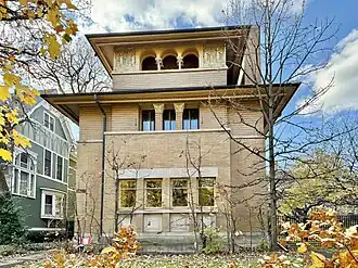 View of the house from the sidewalk. The house has a brick facade with recessed rectangular windows on the first two stories. There is a protruding eave above the second floor. The third-story windows are within an arched loggia.