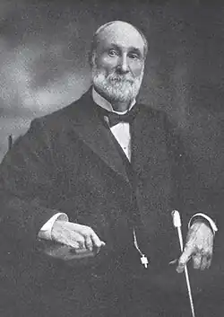 A man with receding gray hair and a gray beard and mustache. He is wearing a black coat, white shirt, and black bow tie
