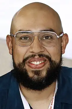 Trejo is smiling directly at the camera in the close-up shot. He is bald with a thick beard, and wearing aviator style wire rimmed eyeglasses.