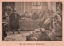 In a room filled with people, a bearded man is standing before a crowned woman who sits on a throne and is surrounded by Catholic priests