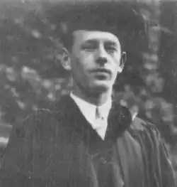 A black and white photo of a clean-shaven man in a graduation uniform , with trees behind him.