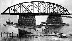 A black and white photograph showing a single truss of a bridge being erected. The steel, semi-circular truss construction is ready and is resting on two pylons erected in the middle of the river and surrounded by scaffolding. Under the bridge a paddle steamer is visible, as well as some wooden temporary causeways and pontoon bridges that allow access to the construction site.