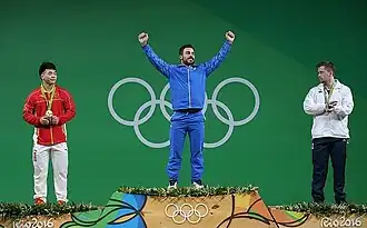 The three medalists for the men's 85kg weightlifting competition standing on the podium. From left to right: Tian Tao of China, Kianoush Rostami of Iran (with his hands raised victoriously), and Denis Ulanov of Ukraine.