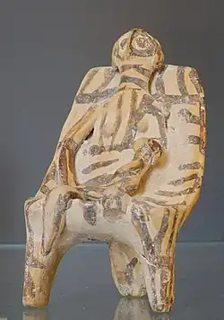 Clay figurine, holding a baby, seated on a throne. The whole thing is fairly crudely made and covered in wavy painted lines.