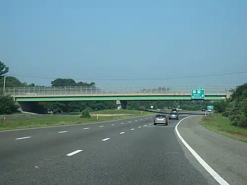 Ground-level view of a three lanes of a divided freeway; a large green and gray overpass bridge and a green exit sign are visible in the distance.