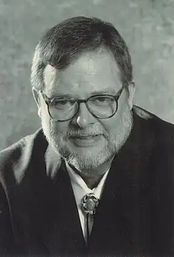 Portrait of Mark Pope, wearing glasses and a dark suit with a bolo tie, facing forward and smiling slightly.