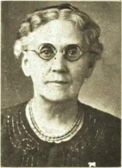 B&W portrait photo of an elderly woman with plaited white hair, wearing glasses, a necklace, and a dark, round-neck jacket.