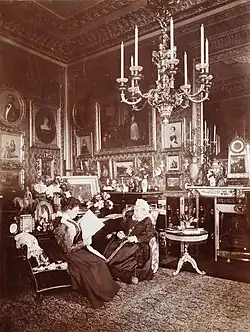 A black and white photograph of an elderly Victoria sat alongside a younger woman (Beatrice) reading a newspaper. The room is ornately decorated, with a number of photographs, paintings and a large chandelier hanging from the ceiling.