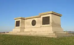 A large monument of off-white stone. Its shape resembles a long, flat pedestal. It sits atop a grassy knoll with a bright blue sky overhead.
