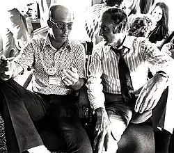 Photograph of two seated men having a conversation in a crowded busy room, the man on the left is giving "the finger" to the camera.