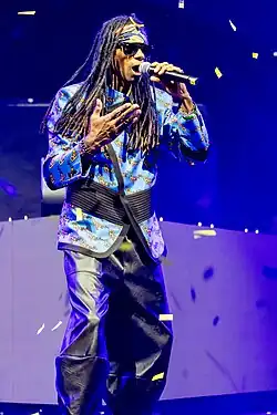 Fab Morvan wearing black leather pants, a colourful, patterned jacket, and black sunglasses, singing into a microphone onstage