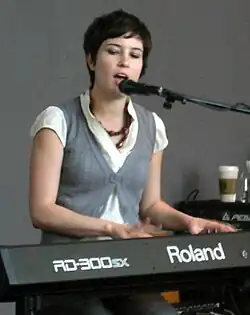Higgins is seated. She sings into a microphone and plays a keyboard instrument. The lettering RD-300SX and Roland are visible across its front.