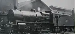 Side-and-front view of a modified version of the N class on shed. The distinguishing feature from normal N class locomotives is the tall cylindrical chimney on the smokebox. A member of the crew is standing next to the tender.