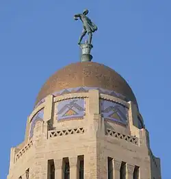 Gold-colored dome atop octagonal stone tower; stylized thunderbirds on sides of tower just below dome; bronze statue of someone sowing seed by hand on top