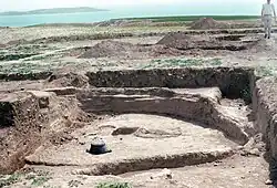 Archaeological site in which we can see an excavation of around twenty square meters of two to three depth meters. The earth is beige. We see the base of a round house. Further on, other similar holes is to see without seeing the contents. On the horizon, there is a range of water.