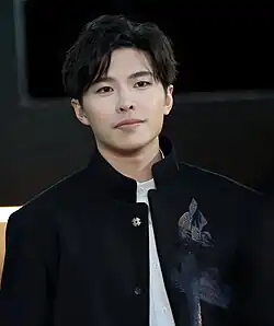 Close up of Yau dressed in a black jacket and smiling while seated on a sofa.