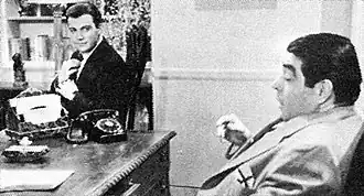 Characters Archie Goodwin (played by Shatner) and Kurt Kasznar are discussing around a business room table. Goodwin is looking to Kasznar while fixing his tie.