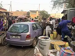 Sacks of grain being sold next to a narrow road with cars and pedestrians.