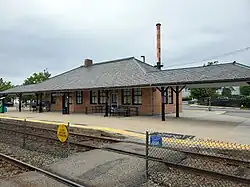 A one-story yellow brick station building next to a rail line. The roofline connects to a canopy which extends both directions along the platform.