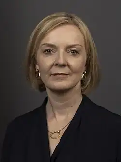 Truss facing frontward, with short blonde hair and dark clothes in front of a grey background.