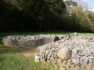 A short dry-stone wall retains boulders to form a cairn. The wall is missing at the front, right section, where the rubble has tumbled out, leaving a (previously covered) orthostat exposed. The wall forms a courtyard at the cromlech's entrance. Flat ground of short grass surrounds the cairn. The background is of shaded trees, mainly in leaf.