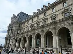 The Louvre museum, a classical building with ornamentation on its facade, rises above a small crowd. Rounded archways line the front of the structure.