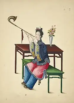 A woman wearing an ao and trousers under an overskirt, she is playing a wind instrument with a curved bell, from 1800.