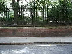 A brick wall topped by metal railings stands at the side of a tarmac road. Behind the wall, grass and trees are visible, growing at the height of the top of the wall.