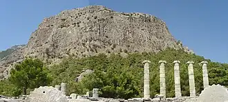 Color photograph of a barren grey mountain, with a small forest at its foothills. Ruined Greek columns can be seen in front of the forest.