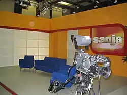 Orange, red and white TV studio, with blue chairs and gray camera