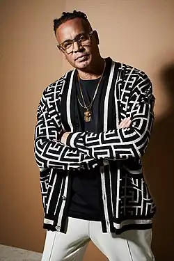 Ray Slijngaard standing with arms crossed, wearing light-coloured pants, a black t-shirt, and a patterned cardigan, looking directly at camera with head slightly tilted to the right